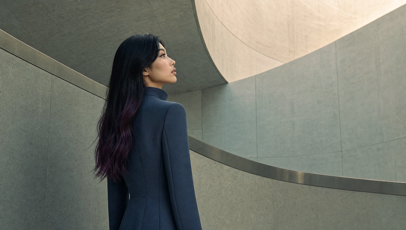 Sora Flame seen from behind in a tailored navy high-neck coat, purple-tipped hair cascading down, gazing upward within a sweeping curved concrete architectural space